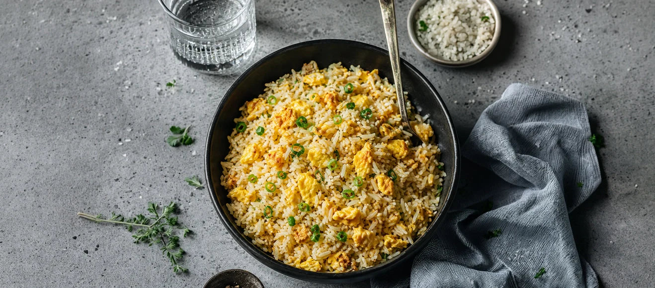 top-down view of a black bowl filled with egg fried rice, garnished with finely chopped green onions. The bowl is set on a textured grey concrete surface next to a glass of water, a small dish of sea salt, a gold spoon, and a crumpled grey linen cloth.