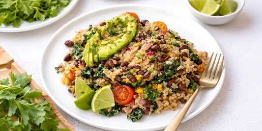 A vibrant Mexican-style rice bowl featuring brown rice mixed with black beans, corn, chopped kale, and cherry tomatoes, topped with fresh avocado slices and cilantro. Served on a white plate with lime wedges and a gold fork on the side.
