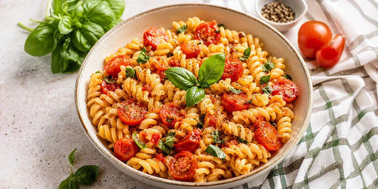 A large ceramic bowl filled with tomato and basil fusilli pasta sits on a light-colored, textured tabletop. 
