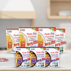 A bulk display of Pasta Zing pouches arranged on a white kitchen countertop with a blurred kitchen background. The back row contains four Spaghetti (orange) pouches, and the front row features three Farfalle (purple) pouches. Every pouch displays the "14g Protein" badge and the quick "1 Minute" microwave cook time, emphasizing the product’s convenience for meal prepping or large families.